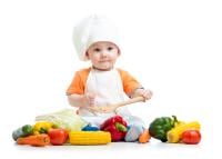 Young boy wearing chefs hat and apron, sat surrounded by some fresh vegetables 