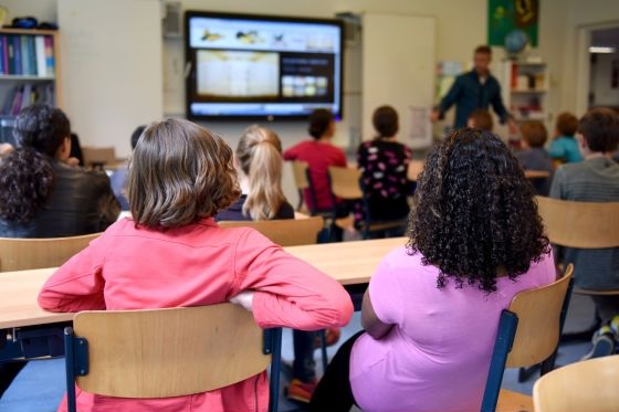 The back of two students, sitting in a classroom, full of children.