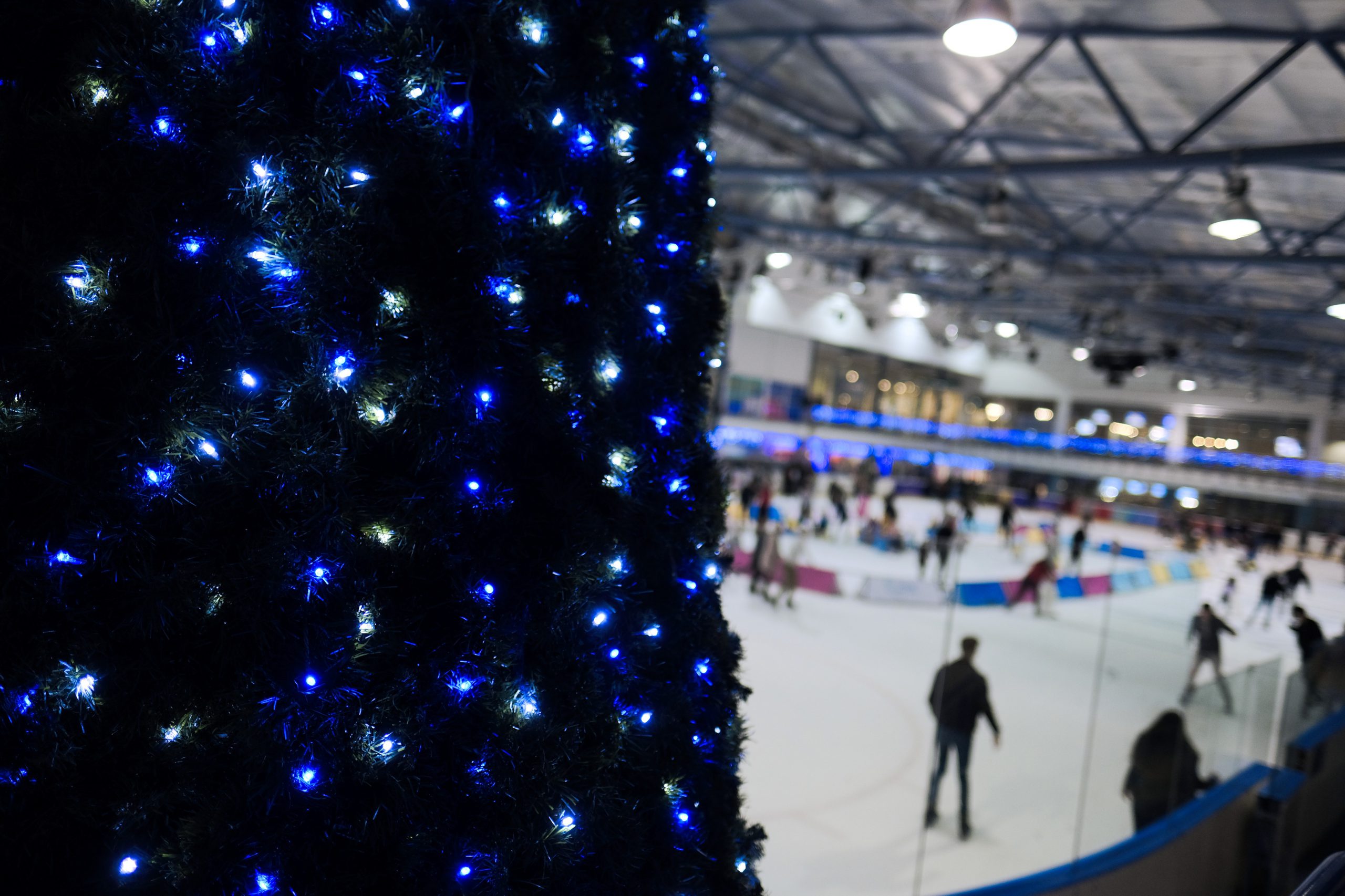 An indoor ice rink filled with people skating, with a large festive decoration covered in blue and white lights in the foreground and illuminated viewing areas above.