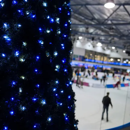 An indoor ice rink filled with people skating, with a large festive decoration covered in blue and white lights in the foreground and illuminated viewing areas above.