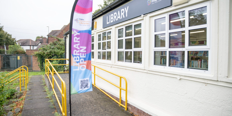 Woodthorpe Library exterior - View of an access ramp leading to a white one story building, windows along the side looking onto the shelves and a multicoloured Library Open flag at the foot of the ramp.